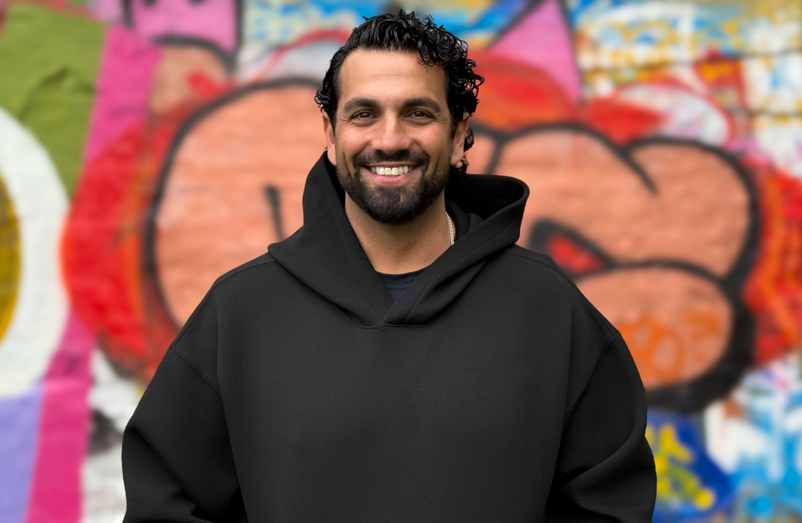 Portrait of a person wearing a black hoodie in front of a colorful graffiti wall.